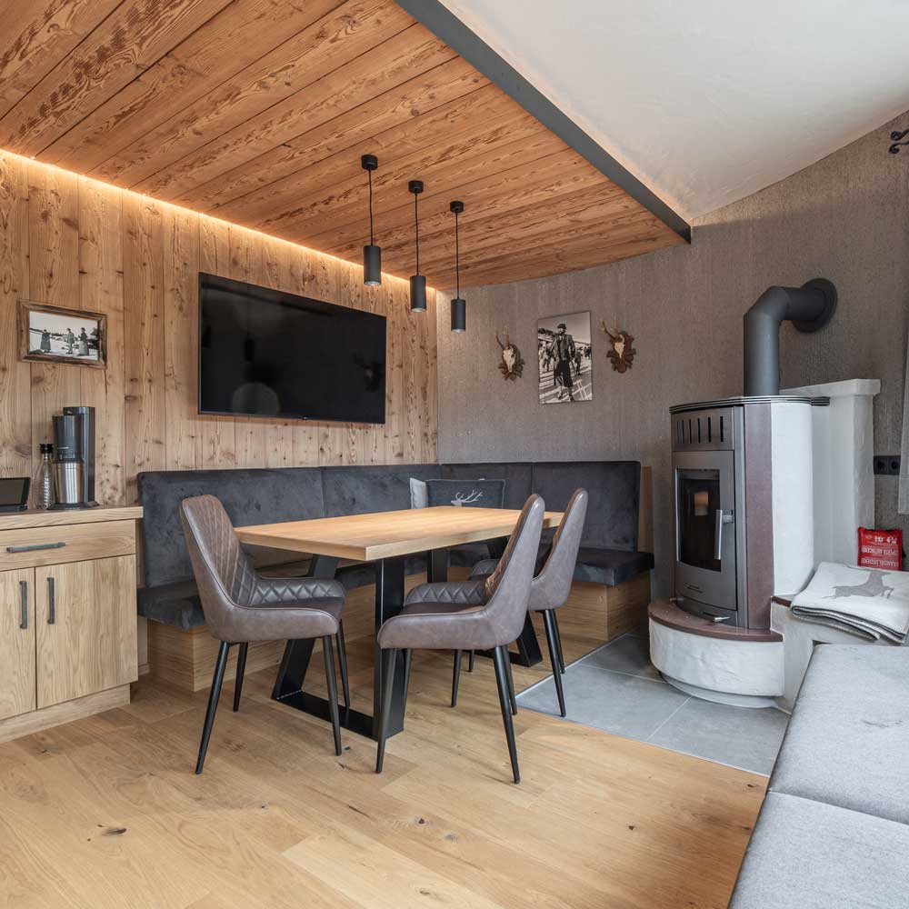 Gemütliches Zimmer im Neuen Apartment Gartenlodge in Ladis mit modernem rustikalem Interieur mit Holzdecke, Eckbank und Esstisch mit grauen Stühlen. Genießen Sie die Wärme eines Holzofens und lebendige Akzente, während Sie auf dem großen Flachbildfernseher an der holzgetäfelten Wand fernsehen.