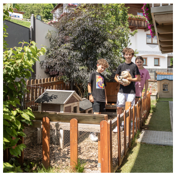 Drei Kinder stehen lächelnd während ihres Familienurlaubs Serfaus-Fiss-Ladis in einem Garten neben einem hölzernen Tiergehege. Eines hält ein Kaninchen. Das Gehege ist zwischen grünen Pflanzen eingebettet, im Hintergrund ist ein mit Blumenkörben geschmücktes Haus zu sehen.