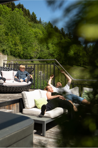Eine Familie genießt einen ruhigen Nachmittag in der Naturlodge am Waldesrand in Ladis und entspannt auf einer sonnenbeschienenen Terrasse mitten im Grünen. Ein Kind sitzt in einem runden Stuhl, während zwei Erwachsene auf gepolsterten Sitzen faulenzen, im Hintergrund eingerahmt von Bäumen und Büschen.