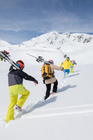Eine Gruppe von vier Personen in bunter Skiausrüstung erklimmt einen schneebedeckten Hügel. Jeder trägt Skier über den Schultern. Im Hintergrund sind schneebedeckte Berge und ein klarer blauer Himmel zu sehen.