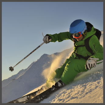 Ein Skifahrer in leuchtend grünem Outfit und blauem Helm fährt eine scharfe Kurve auf einem schneebedeckten Hang. Sonnenlicht erhellt die Szene und lässt den Schnee hinter ihnen aufspritzen. Im Hintergrund sind Berggipfel unter einem klaren blauen Himmel zu sehen.