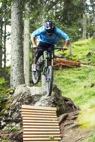 Ein Mountainbiker in blauem Trikot und Helm fährt in einem Waldgebiet eine schräge Holzrampe hinunter. Die Szene ist von üppigen grünen Bäumen und Pflanzen geprägt und der Biker konzentriert sich darauf, den Weg zu bewältigen.
