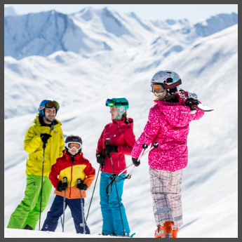 Eine Gruppe von vier Personen, darunter ein Kind, ist in farbenfroher Skiausrüstung gekleidet, lächelt und hält Skistöcke auf einem schneebedeckten Berg. Im Hintergrund sind schneebedeckte Gipfel unter einem klaren blauen Himmel zu sehen.