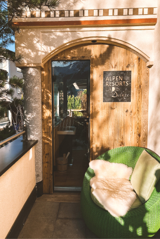 Eine rustikale Holztür mit der Aufschrift „Alpen Resorts“ führt in ein gemütliches Zimmer. Auf der sonnendurchfluteten Terrasse steht ein runder grüner Stuhl mit einer weißen Felldecke. In der hohen Glasscheibe der Tür spiegeln sich Bäume und Berge.