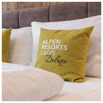 Eine Nahaufnahme eines Bettes mit gepolstertem Kopfteil und weißer Bettwäsche hebt zwei gelbe Kissen hervor, auf denen in klarer Schrift „Alpenresorts Ladis Deluxe“ steht. Der ruhige Hintergrund einer Holzwand vervollständigt die einladende Szene.
