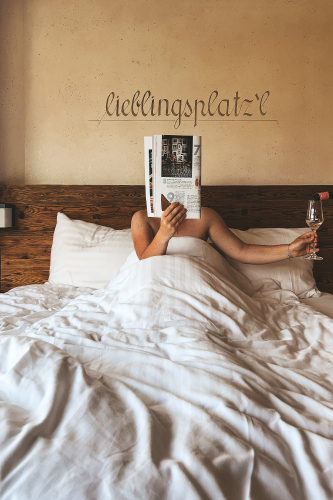 Eine Person liegt im Bett, in ein weißes Laken gehüllt, nippt an einem Wein und blättert in einer Zeitschrift. Das Wort „Lieblingsplatzl“ ziert die beige Wand über dem hölzernen Kopfteil des Bettes und weckt Erinnerungen an einen Urlaub in Ladis. Es ist eine behagliche und entspannte Umgebung.