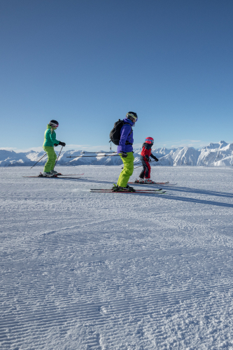 Drei Skifahrer gleiten unter einem klaren blauen Himmel über einen schneebedeckten Berghang und genießen ihren Aktivurlaub in Serfaus-Fiss-Ladis. In heller Winterkleidung, mit Helmen und Skistöcken, schwelgen sie in den schneebedeckten Gipfeln dieses atemberaubenden Reiseziels, das sich sowohl für Sommer- als auch für Winterabenteuer eignet.