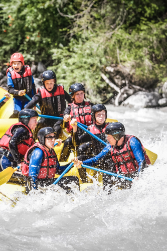 Eine Gruppe Abenteurer genießt einen Aktivurlaub in Serfaus-Fiss-Ladis und bezwingt mit Helmen und Schwimmwesten einen reißenden Fluss. Sie paddeln durch Stromschnellen und genießen den Nervenkitzel, während das Wasser um sie herumspritzt, umgeben von Bäumen und Felsen – eine perfekte Abwechslung sowohl im Sommer als auch im Winter.
