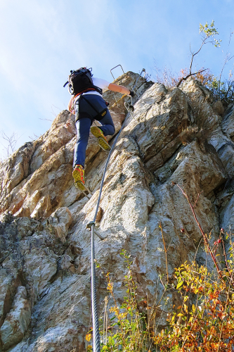 Eine Person mit Rucksack genießt Aktivurlaub in Serfaus-Fiss-Ladis und erklimmt mithilfe eines gesicherten Seils eine Felswand. Der Kletterer, ausgerüstet mit Helm und festem Schuhwerk, steigt unter dem klaren blauen Himmel auf, während die Vegetation den Fuß und den Gipfel dieses Ganzjahres-Abenteuerziels säumt.