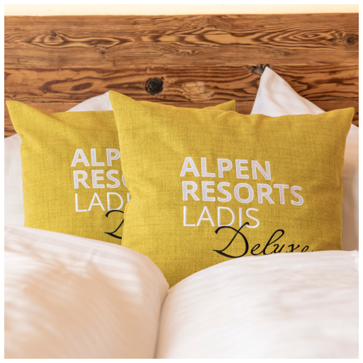 Zwei gelbe Zierkissen mit dem weißen Text „Alpen Resorts Ladis Deluxe“ liegen auf einem Bett mit weißer Bettwäsche. Das hölzerne Kopfteil verleiht der gemütlichen Umgebung eine rustikale Note, perfekt zum Entspannen nach 7 Genussskitagen inkl. 6 Tage Skipass voller alpiner Abenteuer.