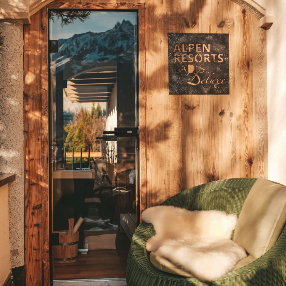 Eine gemütliche Holztür mit Glasscheibe spiegelt Berge und einen Balkon im Freien wider. Auf einem rustikalen Schild steht „Alpen Resorts Lar... Deluxe“. In der Nähe entdecken Sie die private Außensauna in Ladis. Neben der Tür steht ein grüner, gepolsterter Stuhl mit Fellüberwurf, umgeben von gesprenkeltem Sonnenlicht.