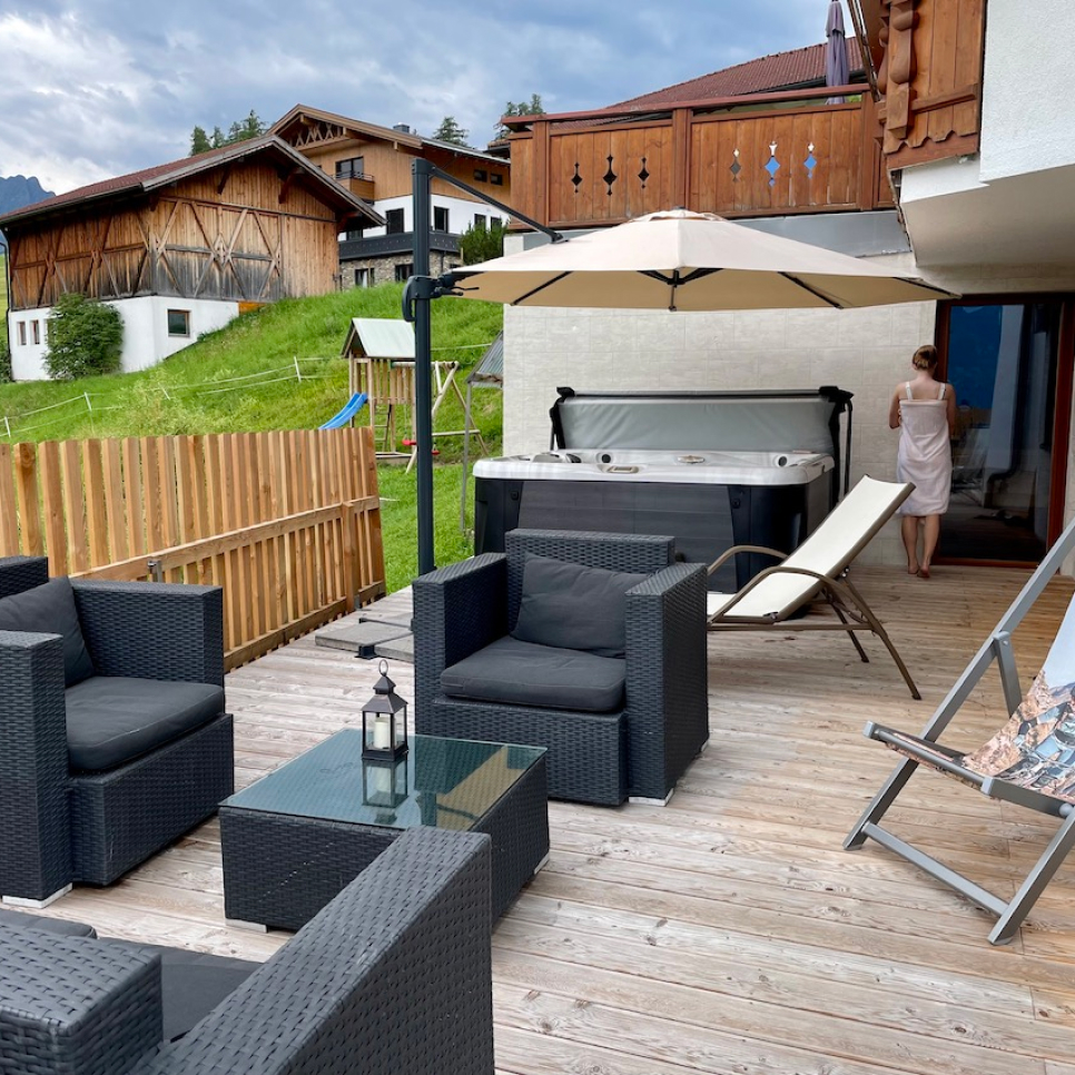 Die Terrasse des Edelweiss Chalet in Ladis ist mit schwarzen Korbmöbeln, Stühlen und einem Glastisch mit Laterne ausgestattet. Genießen Sie einen Whirlpool und eine Sonnenliege unter einem Sonnenschirm. Eine in ein Handtuch gehüllte Person steht an der Tür. Holzhäuser und ein grasbewachsener Hügel verschönern diesen privaten Rückzugsort im Gartenspa.