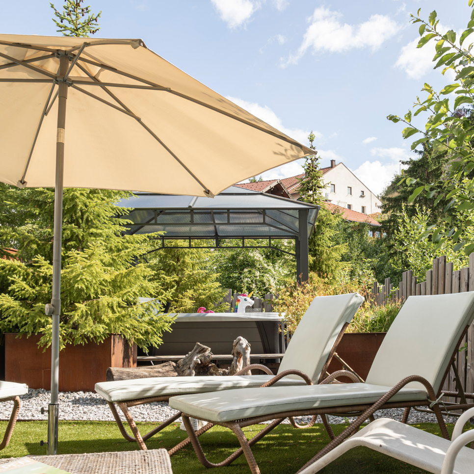Außenterrasse mit weißen Liegestühlen unter einem beigen Sonnenschirm, auf einer Rasenfläche im Garten Spa mit Whirlpool im Laudegghof. Im Hintergrund ist ein kleiner Whirlpool zu sehen, umgeben von üppigen grünen Bäumen. Unter einem klaren Himmel sind auch ein Holzzaun und Dächer von nahe gelegenen Gebäuden zu sehen.
