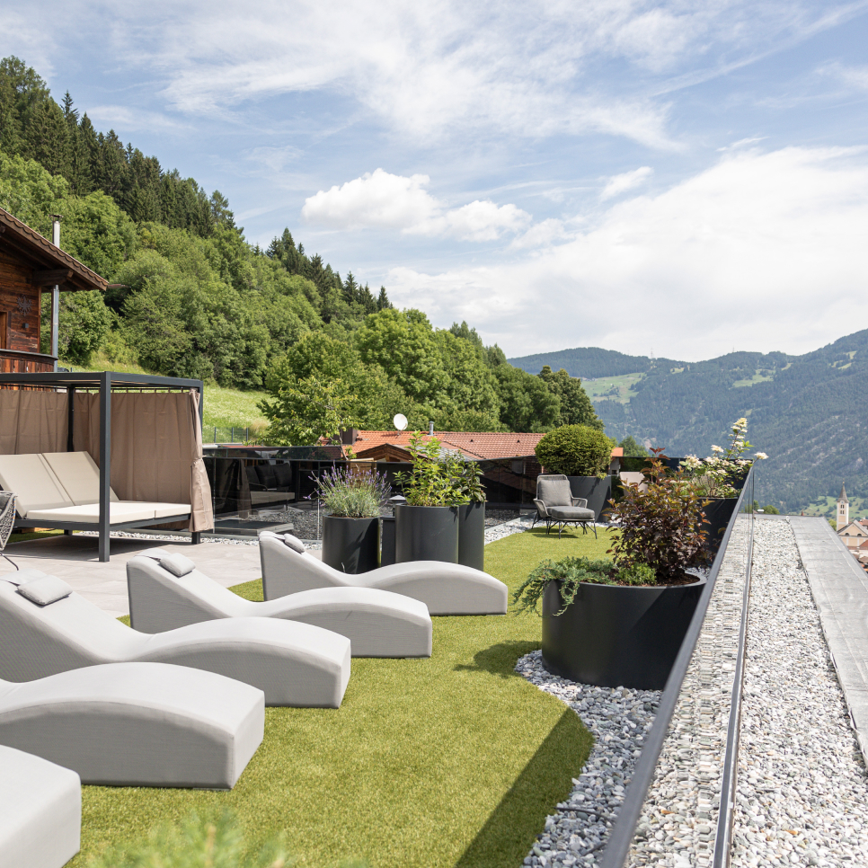 Neue Panoramaterrasse mit Ausblick auf die Bergwelt, ausgestattet mit modernen Liegestühlen und Topfpflanzen. Genießen Sie die malerische Aussicht auf sanfte grüne Hügel und bewaldete Berge unter einem teilweise bewölkten Himmel, alles von Steinkies umgeben.