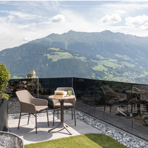 Eine malerische neue Panoramaterrasse mit zwei Stühlen und einem kleinen Tisch vor der Kulisse üppiger, grüner Berge und eines wolkigen Himmels. Der Bereich ist von einem Glasgeländer umgeben und bietet einen unvergleichlichen Ausblick auf die Bergwelt.