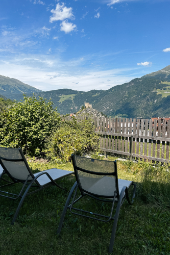 Zwei Liegestühle auf Grasboden blicken auf eine malerische Berglandschaft. Ein Holzzaun verläuft am Rand entlang, in der Nähe befindet sich dichtes Grün. Im Hintergrund sind Hügel und ein Gebäude unter einem blauen Himmel mit vereinzelten Wolken zu sehen.