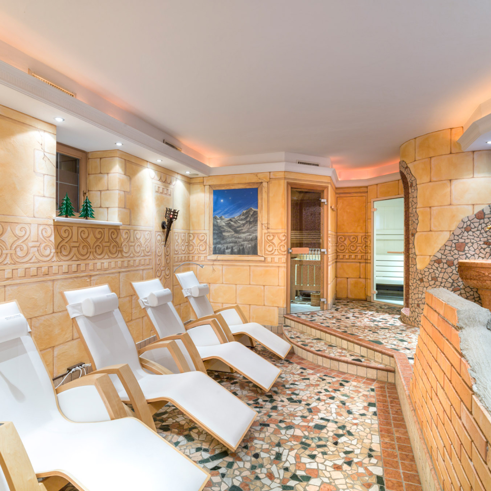 Ein gemütlicher Spa-Raum im Wellnessbereich im Laudegghof verfügt über fünf weiße Liegestühle mit Holzrahmen, Mosaikfliesenboden und warmen, beigen Wänden. Ein malerisches Berggemälde ziert die Wand neben kleinen dekorativen Bäumen und einer charmanten Backsteinstruktur.