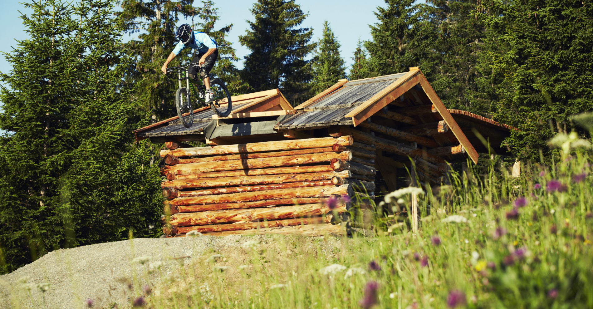 Inmitten der malerischen Landschaft von Serfaus-Fiss-Ladis navigiert ein Mountainbiker gekonnt über ein Holzhüttendach, umgeben von Bäumen und leuchtenden Wildblumen. Er trägt aus Sicherheitsgründen einen Helm und verkörpert den Nervenkitzel dieses unvergesslichen Bikeurlaubs.