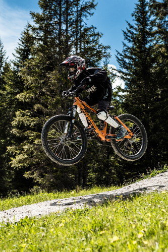 Ein Mountainbiker in voller Montur, inklusive Helm, schwebt in der Luft, nachdem er während seines Bikeurlaubs in Serfaus-Fiss-Ladis von einer Sandrampe gesprungen ist. Das orange-schwarze Bike gleitet vor der Kulisse von Kiefern und einem klaren blauen Himmel dahin, und am Landeplatz wartet grünes Gras.