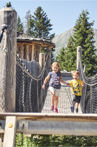 Zwei Kinder laufen fröhlich über eine Hängebrücke im bergigen Familienurlaub in Serfaus Fiss Ladis. Das Mädchen trägt ein graues Hemd und der Junge ein gelbes, während Bäume und eine Holzkonstruktion stolz gegen den klaren Himmel ragen.