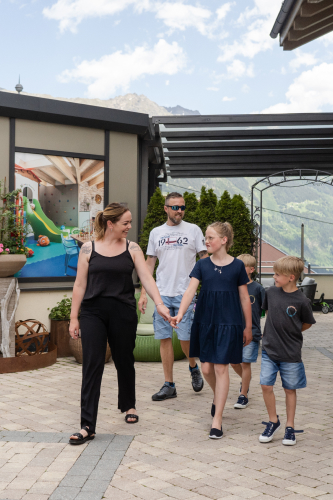 Eine vierköpfige Familie genießt ihren Familienurlaub in Serfaus Fiss Ladis und spaziert einen gepflasterten Außenweg entlang. Die Erwachsenen in Freizeitkleidung führen die Kinder, die einen Elternteil an der Hand halten. Üppige Vegetation und majestätische Berge bestimmen die Szenerie unter einem teilweise bewölkten Himmel.