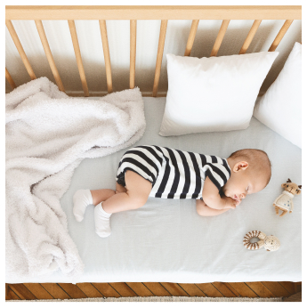 Ein Baby in einem schwarz-weiß gestreiften Strampler schläft friedlich auf der Seite in einem Kinderbett, umgeben von kuscheliger weißer Bettwäsche, einer flauschigen Decke und zwei Puppen. Die hellen Holzgitter erinnern sanft an ruhige Nächte, in denen man vom Familienurlaub in Serfaus Fiss Ladis träumte.