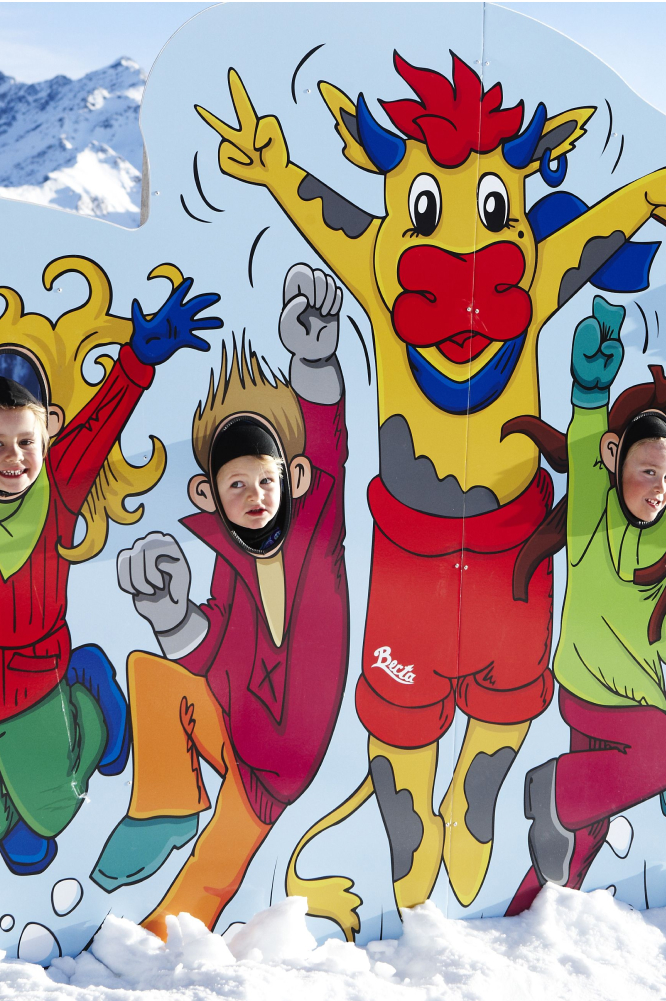 Drei Kinder posieren während ihres Familienurlaubs in Serfaus Fiss Ladis fröhlich mit ihren Gesichtern in Ausschnitten eines farbenfrohen Cartoon-Figuren-Aufstellers – die Figuren ähneln einem Stier und Menschen in Winterkleidung vor einer malerischen Kulisse aus schneebedeckten Bergen.