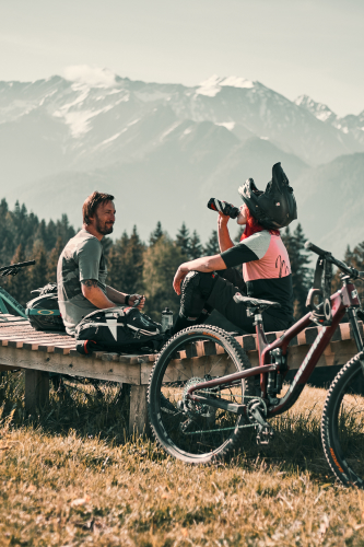 Zwei Personen in Radsportkleidung entspannen auf einer Bank, ihre Mountainbikes stehen daneben und genießen ihren Bikeurlaub in Serfaus-Fiss-Ladis. Einer nippt an einer Flasche, während schneebedeckte Berge und Bäume den klaren Himmel im Hintergrund einrahmen.