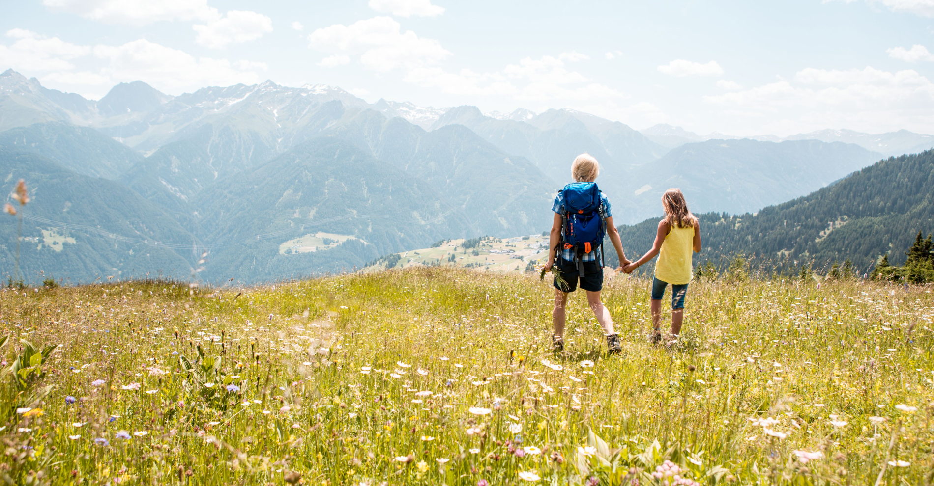 Ein Erwachsener und ein Kind halten sich an den Händen und wandern durch eine blühende Wiese mit Wildblumen. Das ist ein Bild, das die Essenz eines Sommerurlaubs in Serfaus-Fiss-Ladis einfängt. Sie blicken auf die Berge in der Ferne unter einem teilweise bewölkten Himmel, was auf einen warmen Tag schließen lässt. Der Erwachsene trägt einen Rucksack und Wanderstöcke.