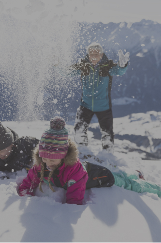 Ein Kind in einer rosa Jacke und einer bunten Mütze liegt lächelnd im Schnee. Eine andere Person in einer blauen Jacke steht dahinter und wirft spielerisch Schnee in die frische Alpenluft. Schneebedeckte Berge rahmen diese fröhliche Szene ein, die an Abenteuer in den Alpenresorts erinnert.
