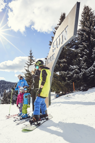 Eine Gruppe Kinder in bunter Skiausrüstung steht auf einem schneebedeckten Hang unter einem großen Schild mit der Aufschrift „SUN PARK“. Sie genießen ihren Winterurlaub in Serfaus Fiss Ladis, tragen Helme, die Sonne scheint hell gegen den klaren blauen Himmel, Bäume rahmen die malerische Szene ein.