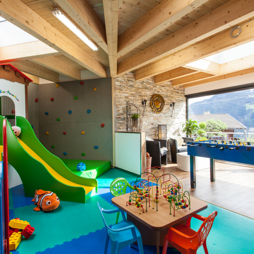 Ein helles Spielzimmer in Mimis Kinderspielwelt im Alpenapart in Ladis verfügt über eine Kletterwand, eine Rutsche und einen Tischfußballtisch. Bunte Stühle stehen um einen Tisch mit Perlenlabyrinthen herum, während große Fenster einen malerischen Blick auf die Berge ermöglichen.