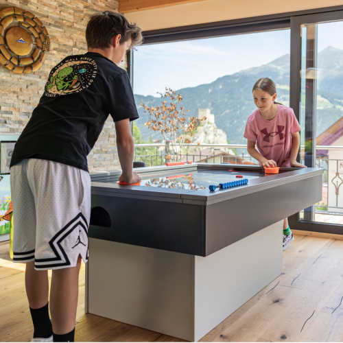 In Mimis Kinderspielwelt im Alpenapart in Ladis spielen zwei Kinder an einem großen Tisch mit Schlägern ein Spiel. Der Junge trägt ein schwarzes Shirt und weiße Shorts, das Mädchen ist in Rosa gekleidet. Sie genießen den Blick auf die Berge durch die Glaswand und stehen auf einem Holzboden vor einer mit Steinen gestalteten Wand.