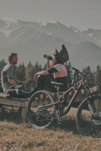 Zwei Mountainbiker ruhen sich auf einer Holzbank in einem Alpenresort aus, umgeben von Bäumen und schneebedeckten Bergen in der Ferne. Eine Person trägt einen Helm und trinkt aus einer Flasche, während die andere zusieht. Ihre Fahrräder lehnen in der Nähe.
