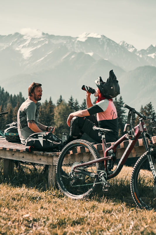 Zwei Personen machen während ihres Sommerurlaubs in Serfaus-Fiss-Ladis eine Pause auf einer Bank mit Mountainbikes in der Nähe. Einer trägt einen Helm und trinkt aus einer Flasche, während der andere sich ausruht. Beide blicken im Hintergrund auf schneebedeckte Berge, was eine friedliche und malerische Aussicht bietet.