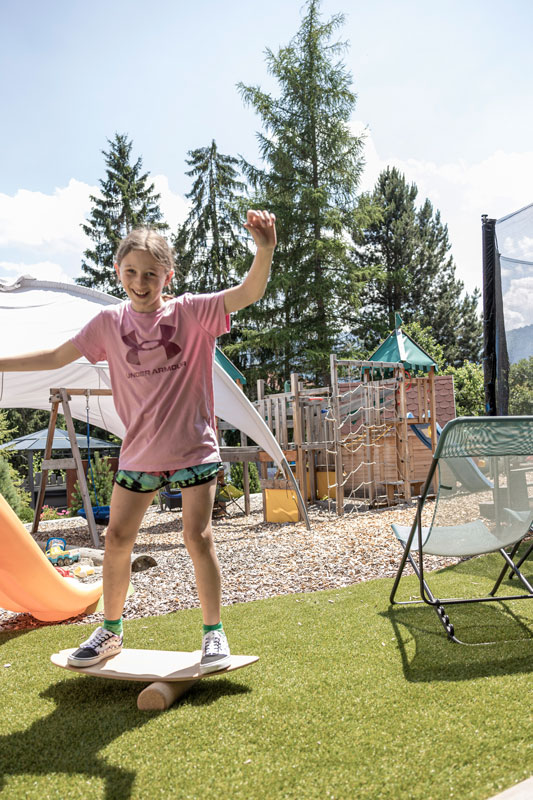 Ein Kind in einem rosa Hemd balanciert auf einem Holzbrett auf einer zylindrischen Rolle in einem Hinterhof, der an einen Sommerurlaub in Serfaus-Fiss-Ladis erinnert. Eine Rutsche, ein Klettergerüst und ein Trampolin stehen in der Nähe, umgeben von Bäumen unter dem klaren Himmel.