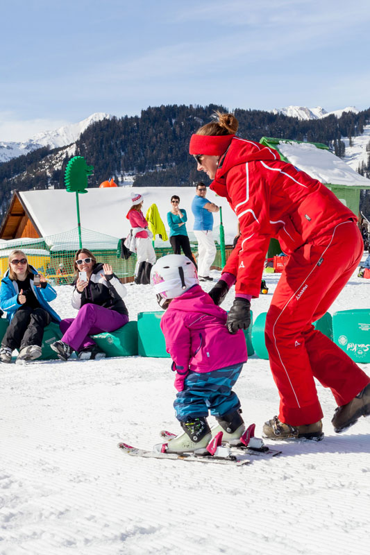 Ein Skilehrer im roten Skianzug führt ein Kleinkind in rosa Skiausrüstung während eines Winterurlaubs in Serfaus Fiss Ladis die verschneiten Hänge hinunter. Im Hintergrund genießen andere Skifahrer und Zuschauer die atemberaubende Bergsicht unter einem klaren Himmel.