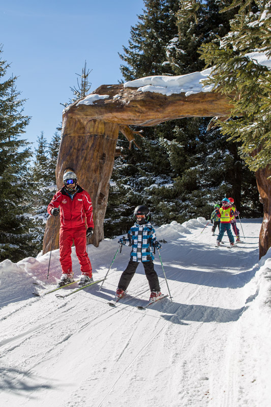 Eine Gruppe Skifahrer, darunter zwei Erwachsene und ein Kind, genießt ihren Winterurlaub in Serfaus Fiss Ladis. In farbenfroher Ausrüstung gleiten sie unter einem Holzbogen auf einer verschneiten Piste entlang, umgeben von schneebedeckten Bäumen unter einem klaren blauen Himmel.