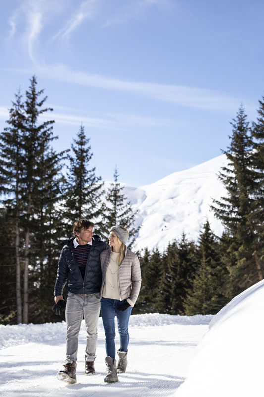 Ein Paar spaziert Arm in Arm einen verschneiten Weg entlang, der von Tannen gesäumt ist, mit einem schneebedeckten Berg im Hintergrund. Warm eingepackt in Jacken und Stiefeln genießen sie unter dem klaren blauen Himmel ihren Winterurlaub in Serfaus Fiss Ladis.