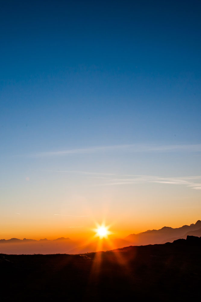 Sonnenaufgang über einer Berglandschaft in Serfaus-Fiss-Ladis, mit einem klaren, abgestuften Himmel, der am Horizont von tiefblau zu orange wechselt. Die Sonne ist teilweise sichtbar, wirft einen warmen Schein auf die entfernten Gipfel und schafft eine heitere Atmosphäre – perfekt für ein Bikeurlaub-Abenteuer am frühen Morgen.