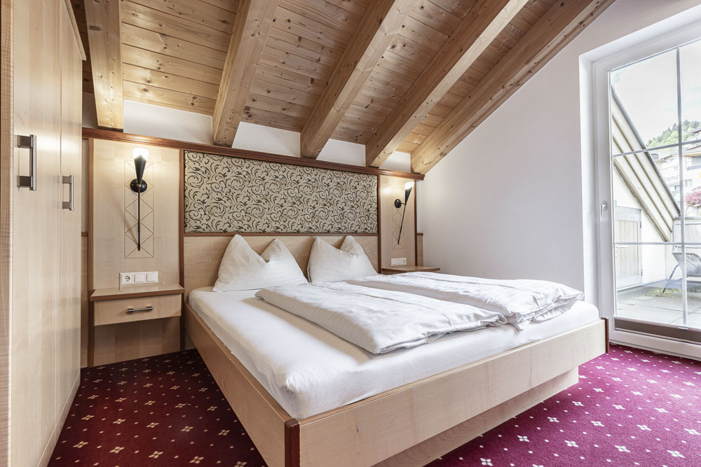 Ein gemütliches Schlafzimmer im Apartment Abendsonne im Alpenapart in Ladis mit schräger Holzdecke und einem Doppelbett mit weißer Bettwäsche und dekorativen Kissen. Wandlampen, gemusterte Tapeten und ein roter Teppich sorgen für Charme, während natürliches Licht durch ein großes Fenster strömt.