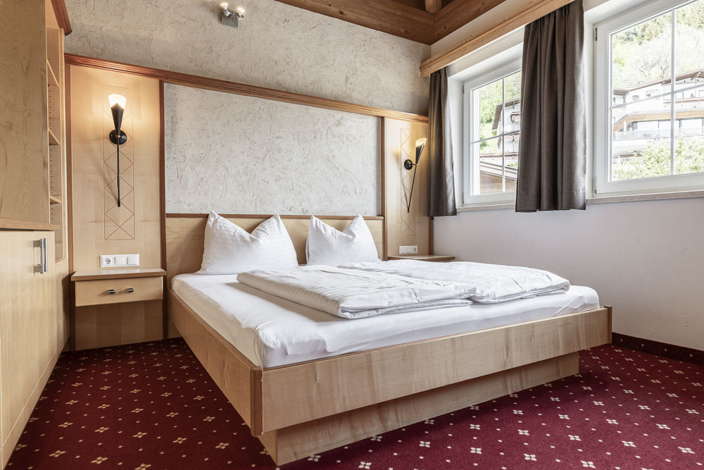 Modernes Apartment-Schlafzimmer im Abendsonne im Alpenapart in Ladis mit Holzbett, weißer Bettwäsche und zwei Wandleuchten. Ein roter Teppich mit weißen Mustern verstärkt den Charme. Dunkle Vorhänge rahmen das Fenster auf der rechten Seite ein und lassen natürliches Licht in diesen einfachen, aber einladenden Raum.