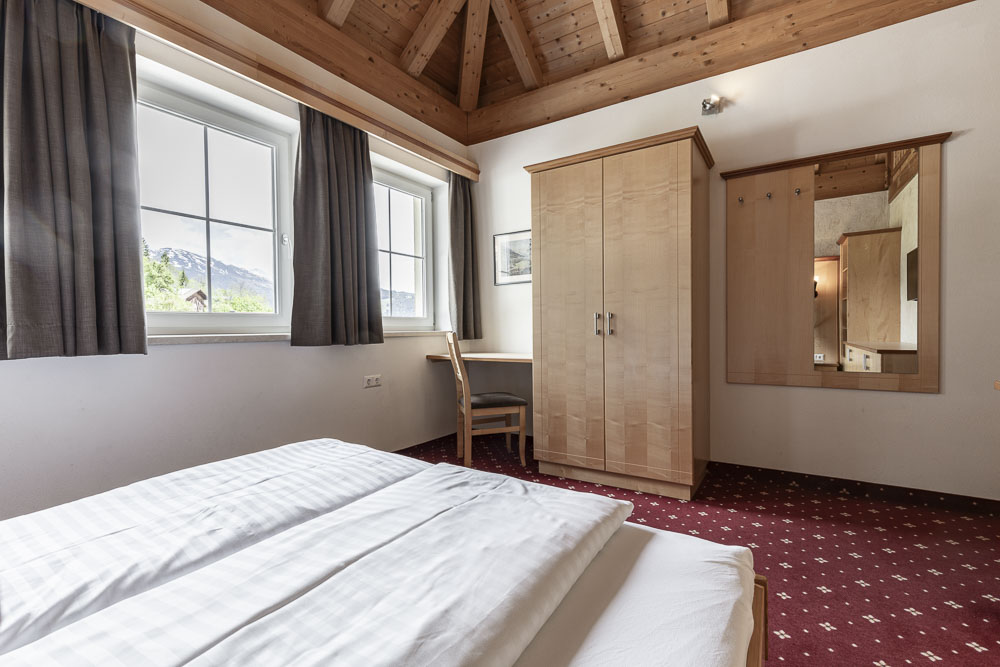 Ein gemütliches Schlafzimmer im charmanten Apartment Abendsonne im Alpenapart in Ladis mit schräger Holzdecke. Es verfügt über ein ordentlich gemachtes Bett, einen Holzschrank, einen großen Spiegel und einen Schreibtisch mit Stuhl am Fenster. Der rote Teppich ergänzt den atemberaubenden Bergblick.