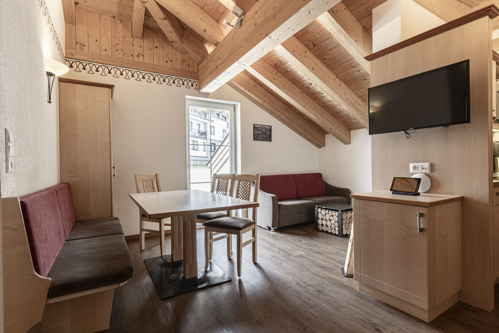 Erleben Sie den gemütlichen Charme des Apartments Abendsonne im Alpenapart in Ladis, dessen Holzbalken die Decke schmücken. Genießen Sie einen Esstisch, eine kastanienbraune Plüschbank und ein Sofa unter dem hellen Tageslicht, das durch das Fenster hereinfällt. Ein an der Wand montierter Fernseher und ein Schrank vervollständigen diesen einladenden Rückzugsort.