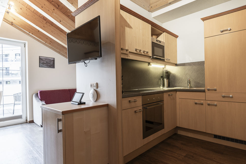 Die gemütliche Küche im Apartment Abendsonne im Alpenapart in Ladis verfügt über helle Holzschränke, einen eingebauten Ofen und einen Elektroherd. Ein an der Wand montierter Fernseher steht gegenüber einem beigefarbenen Sofa. Der Raum verfügt über eine schräge Holzdecke und eine Glastür, die auf einen Balkon führt und natürliches Licht hereinlässt.