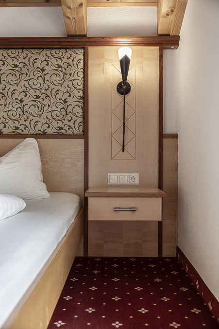 Eine gemütliche Ecke im Apartment Abendsonne im Alpenapart in Ladis verfügt über ein Einzelbett und eine kunstvolle Wandgestaltung. Eine moderne Lampe und Steckdosen schmücken die Wand, während der rot gemusterte Teppich die neutralen Töne und Holzakzente ergänzt und einen warmen alpinen Rückzugsort schafft.