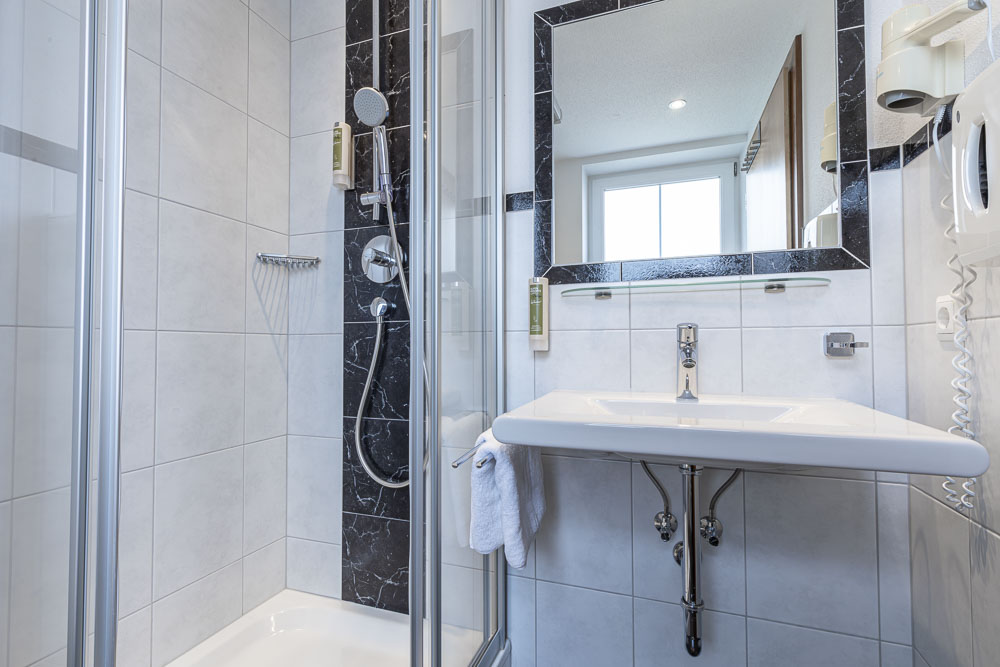 Modernes Badezimmer im Apartment für 4-6 Personen im Alpenapart in Ladis mit einer verglasten Dusche auf der linken Seite mit Regenduschkopf. Über einem weißen Waschbecken hängt ein großer Spiegel, an dessen Rand ein Handtuch hängt. Die Wände sind elegant in Weiß und Schwarz gefliest und reflektieren das natürliche Licht des Fensters.