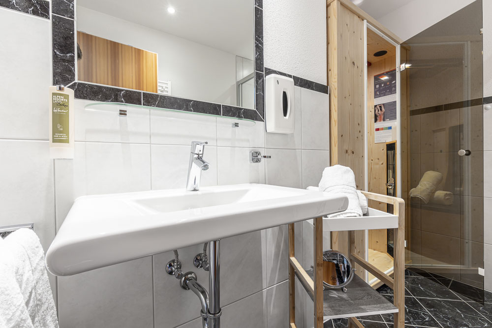 Das moderne Badezimmer im Apartment für 4-6 Personen im Alpenapart in Ladis verfügt über ein weißes Waschbecken, einen wandmontierten Seifenspender und einen großen Spiegel. Ein Holzstuhl hält weiße Handtücher bereit. Das elegante Design umfasst eine verglaste Dusche und eine Holztür – perfekt für Ihren Alpenurlaub.