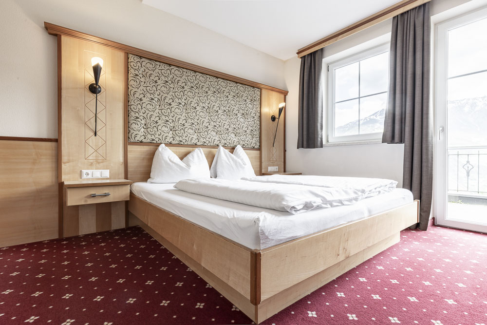 Dieses Hotelzimmer im Alpenapart in Ladis ist ein moderner Rückzugsort in den Alpen und bietet Platz für 4-6 Gäste. Es verfügt über ein großes Bett mit weißer Bettwäsche und Kissen, einen Holzrahmen mit Wandlampen, einen gemusterten roten Teppich, ein dekoratives Kopfteil und Vorhänge, die die malerische Aussicht einrahmen.