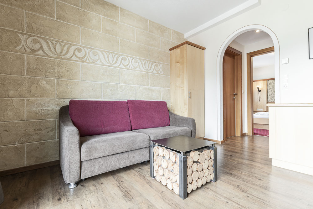 Ein gemütliches Wohnzimmer in unserem Apartment für 4-6 Personen im Alpenapart in Ladis verfügt über ein graues und kastanienbraunes Sofa vor einer Wand mit Steinmuster. Davor steht ein Couchtisch aus gestapelten Holzscheiten. Ein Torbogen führt zu einem Schlafzimmer, alles auf hellem Holzboden und mit modernem und rustikalem Dekor eingerichtet.